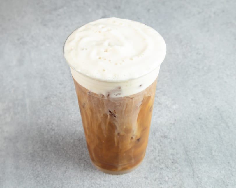 Iced coffee with a thick layer of white foam on top, in a clear plastic cup.