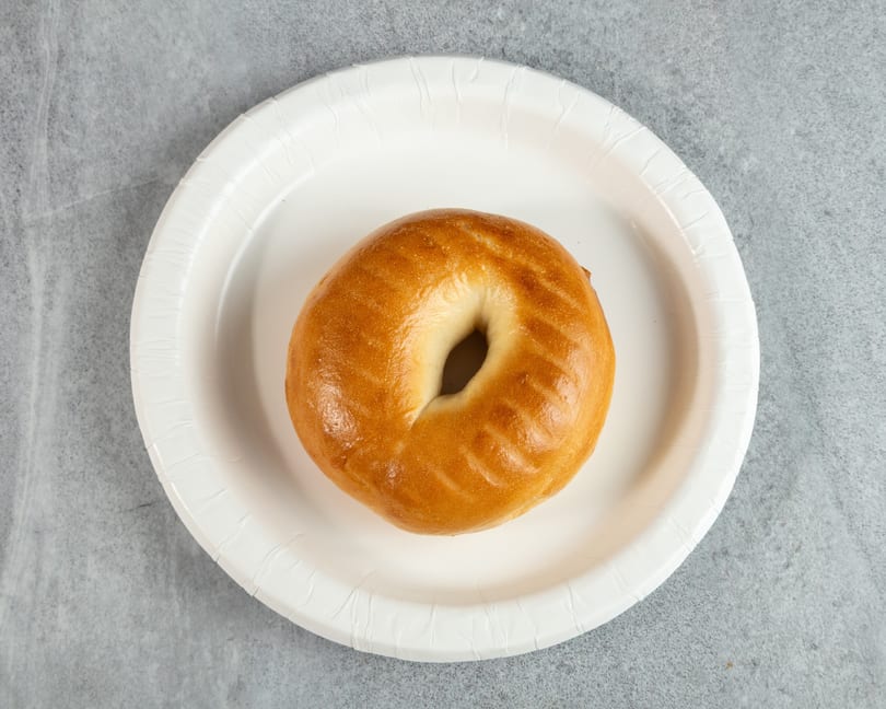 A plain, golden-brown bagel sits centered on a white paper plate against a gray background.