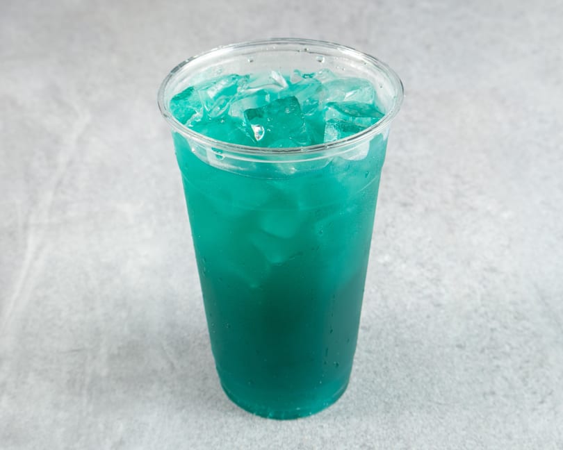 A clear plastic cup filled with a vibrant blue-green drink and ice cubes, on a gray surface.