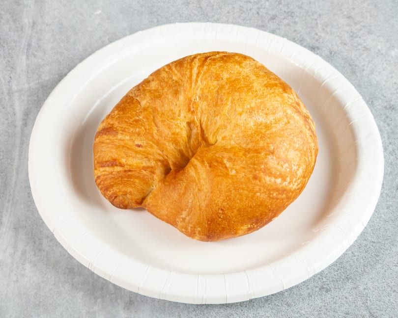 Golden-brown croissant on a white paper plate, centered on a gray textured surface.