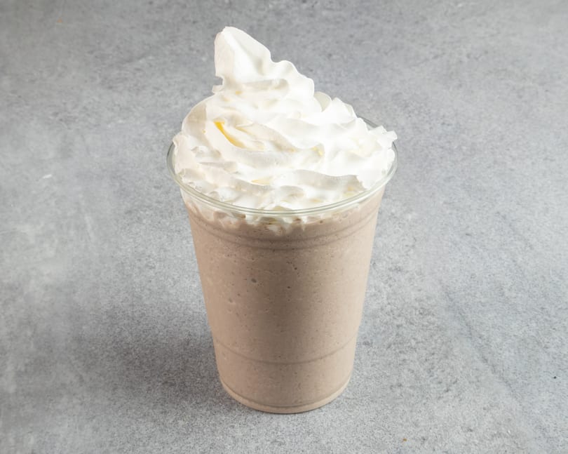 A chocolate milkshake in a clear plastic cup, topped with a generous swirl of white whipped cream.