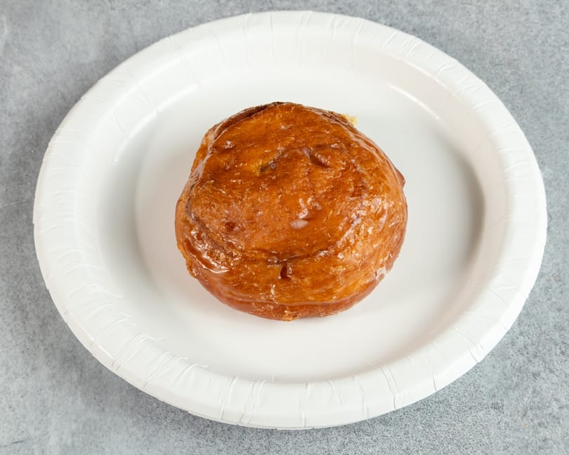 A single glazed apple fritter sits on a white paper plate against a gray background.