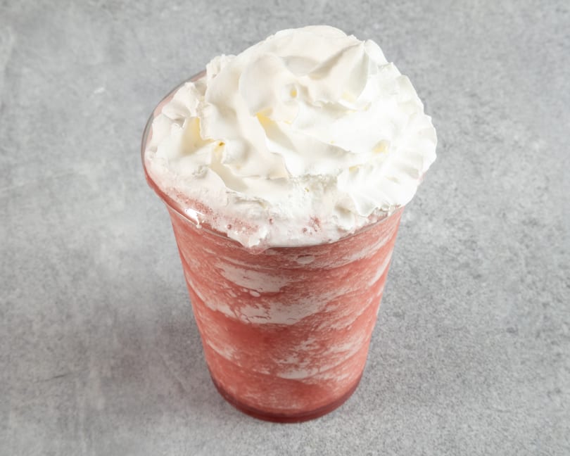Pink frozen drink with whipped cream in a clear plastic cup on a gray surface.