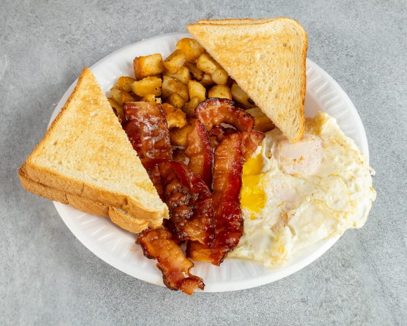 A breakfast platter with crispy bacon, fried eggs, home fries, and buttered toast on a white plate.