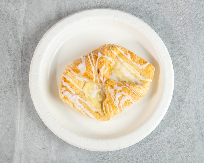 A square cheese danish with white icing drizzle sits on a white paper plate.
