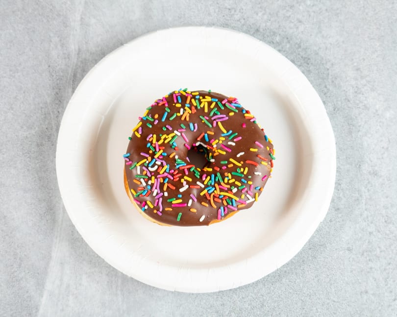 Chocolate frosted donut with colorful sprinkles on a white paper plate.